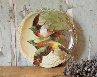 Pode incluir: Um prato de porcelana vintage com um design decorativo de três pássaros em voo. Os pássaros são pintados em tons de verde, marrom, amarelo e vermelho sobre um fundo creme. O prato tem uma borda festonada e um design floral.