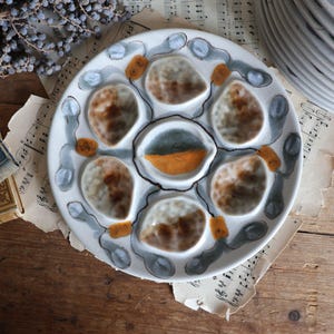Puede incluir: Un plato de ostras de cerámica blanca con seis platos de servicio individuales. El plato está decorado con un diseño de espiral gris y negro y cada plato tiene un glaseado marrón y amarillo.