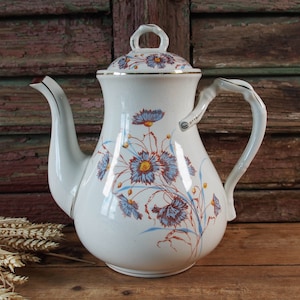 Może przedstawiać: Biały ceramiczny czajnik z kwiatowym wzorem z niebieskimi, czerwonymi i żółtymi kwiatami. Czajnik ma złotą obwódkę wokół pokrywki i uchwytu.