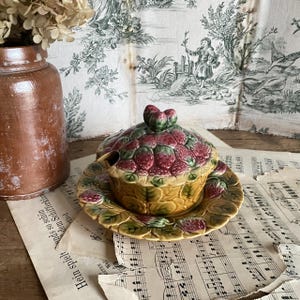 May include: A ceramic strawberry-shaped serving dish with a matching saucer. The dish is yellow with green leaves and a lid covered in red strawberries. A small spoon rests on the side. The background includes sheet music and a brown ceramic jar.