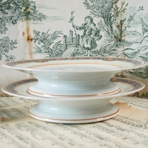 Op de afbeelding: Twee witte porseleinen serveerschalen met een blauw en goud patroon op de rand. De schalen zijn op elkaar gestapeld, waardoor een gelaagd effect ontstaat. De achtergrond is een vintage toile stof met een groen-witte pastorale sc&egrave;ne.