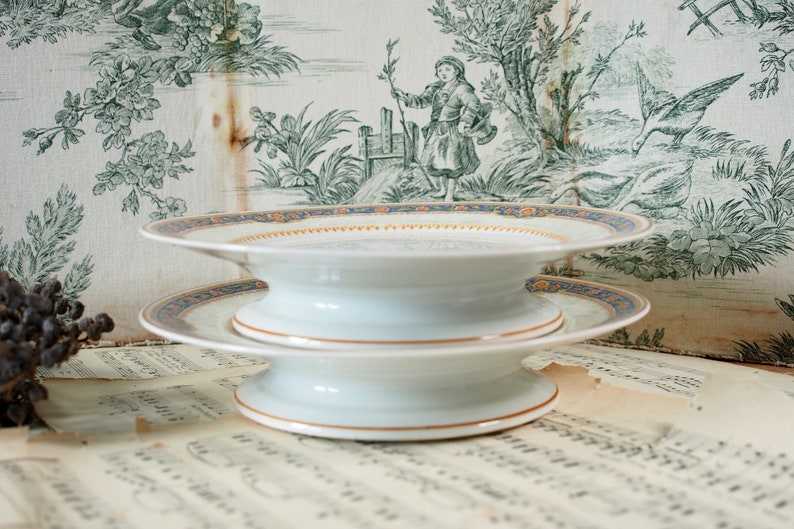 Op de afbeelding: Twee witte porseleinen serveerschalen met een blauw en goud patroon op de rand. De schalen zijn op elkaar gestapeld, waardoor een gelaagd effect ontstaat. De achtergrond is een vintage toile stof met een groen-witte pastorale sc&egrave;ne.
