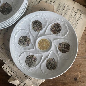 Op de afbeelding: Wit keramisch oesterserveerbord met zes oesterschelpvormige compartimenten, elk met een gedetailleerde oesterillustratie. Een centrale put bevat een citroenschijfje.