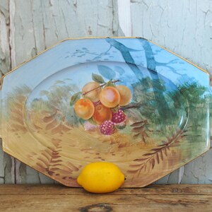 May include: A vintage octagonal serving platter with a hand-painted scene of fruit and foliage. The platter has a light blue background with a gold rim and a yellow lemon in the foreground.