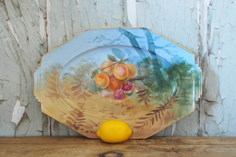 May include: A vintage octagonal serving platter with a hand-painted scene of fruit and foliage. The platter has a light blue background with a gold rim and a yellow lemon in the foreground.