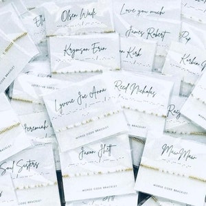 May include: A close-up of several white cards with black text and gold bead bracelets. The cards have names and messages written on them in black ink. The bracelets are made of small gold beads and are arranged on the cards. The cards are on a white surface.