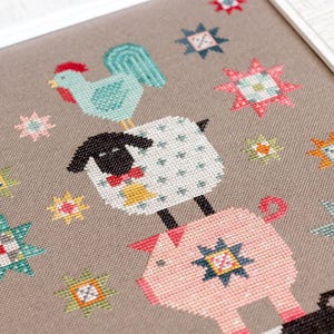 May include: Cross-stitch design showing a teal rooster, a white sheep with a black face, and a pink pig, stacked. The design includes colourful star accents. The artwork is set against a neutral background.