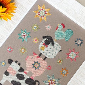 May include: A framed cross-stitch depicting farm animals and starburst patterns. The animals, a cow, pig, sheep, and rooster, are rendered in a playful style. The colour scheme incorporates pink, blue, yellow, and black against a neutral backdrop.