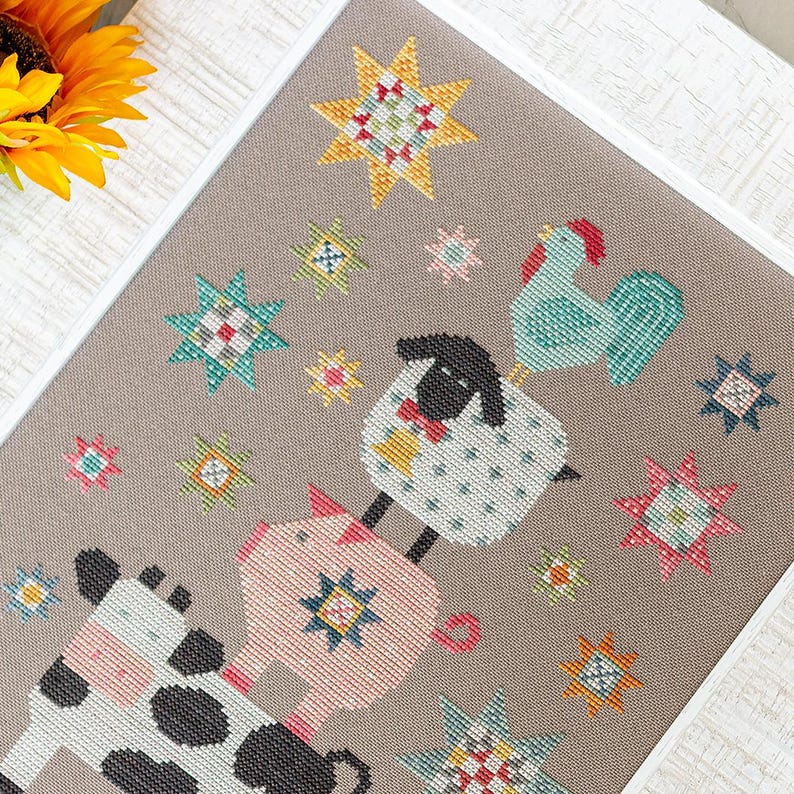 May include: A framed cross-stitch depicting farm animals and starburst patterns. The animals, a cow, pig, sheep, and rooster, are rendered in a playful style. The colour scheme incorporates pink, blue, yellow, and black against a neutral backdrop.