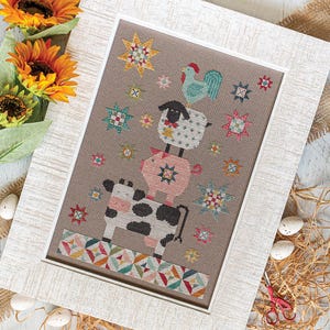 May include: Cross-stitch picture in a white frame, depicting a cow, pig, sheep, and rooster balanced on top of each other. The background is neutral with colourful star and geometric patterns.