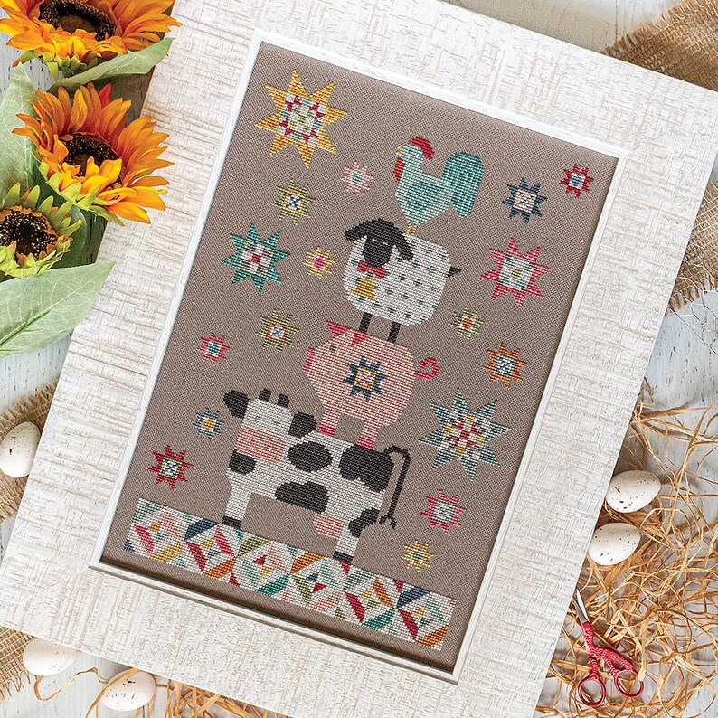 May include: Cross-stitch picture in a white frame, depicting a cow, pig, sheep, and rooster balanced on top of each other. The background is neutral with colourful star and geometric patterns.