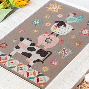 May include: A framed cross-stitch with a farm animal theme. A cow, pig, sheep, and rooster are stacked, with colourful star accents. The artwork is framed in a white border, showcasing a folk art style.