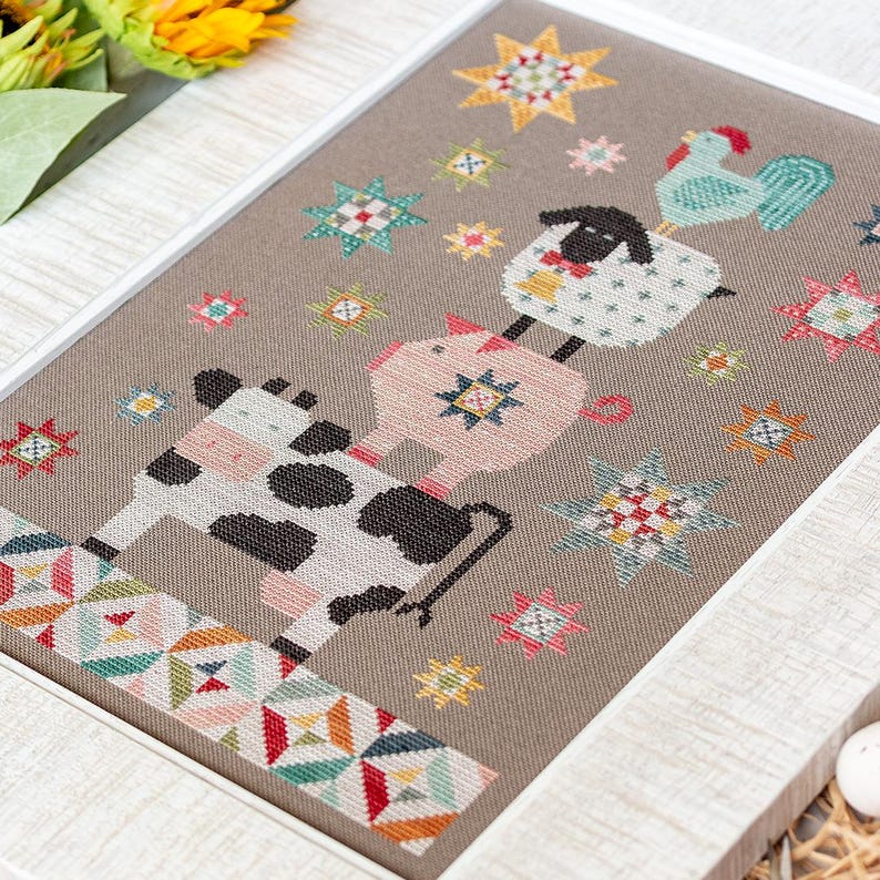 May include: A framed cross-stitch with a farm animal theme. A cow, pig, sheep, and rooster are stacked, with colourful star accents. The artwork is framed in a white border, showcasing a folk art style.