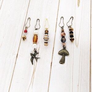 May include: A collection of five decorative stitch markers. Each marker features a safety pin clasp and is adorned with beads in various colors, including red, gold, brown, and black. One marker has a bird charm, and another has a mushroom charm.