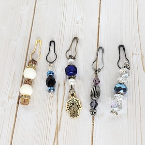 May include: Five beaded stitch markers with safety pin clasps. The markers feature various beads in white, black, blue, and clear, with gold and silver-toned metal accents. One marker has a gold-toned Hamsa charm.