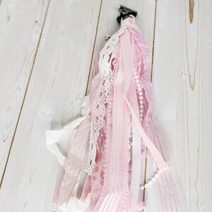 May include: A decorative tassel featuring various shades of pink and white ribbons, lace, and tulle. The tassel is attached to a black clip, and the ribbons are of varying textures and widths. A string of small white beads adds detail.