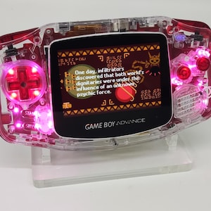 May include: A clear pink Game Boy Advance handheld gaming console with a bright pink backlight. The screen displays a video game with text that reads "One day, infiltrators discovered that both worlds' psychic force..."