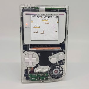 Op de afbeelding: Een transparante Game Boy handheld console met een klassiek Mario spel. Het scherm toont een pixel level met Mario. De interne componenten van de console zijn zichtbaar, en de tekst "GAME BOY" is aanwezig.