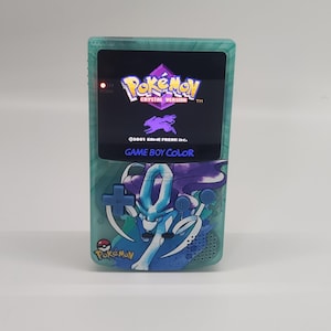 Op de afbeelding: Een turquoise Game Boy Color met een aangepaste frontplaat met een blauw en paars Pokémon-personage. Het scherm toont het titelscherm van de Pokémon Crystal-versie.
