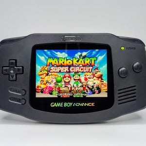 Könnte beinhalten: Eine schwarze Game Boy Advance Handheld-Spielkonsole mit einem leuchtenden Farbbildschirm, auf dem das Spiel Mario Kart Super Circuit angezeigt wird. Der Bildschirm zeigt den Titelbildschirm des Spiels mit einer Gruppe von Charakteren aus der Mario Kart-Serie.