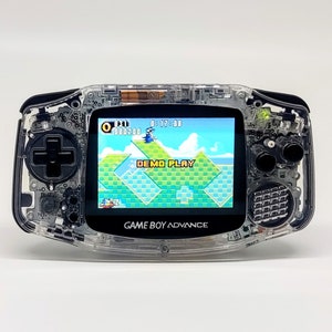 Könnte beinhalten: Eine transparente, durchsichtige Game Boy Advance Handheld-Spielkonsole mit einem schwarzen Steuerkreuz und Knöpfen. Der Bildschirm zeigt ein Videospiel mit dem Text "DEMO PLAY".
