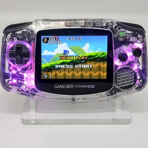 May include: A transparent Game Boy Advance handheld console with purple LED lights. The screen displays a Sonic the Hedgehog game. The console is on a clear stand. The text "GAME BOY ADVANCE" is visible.