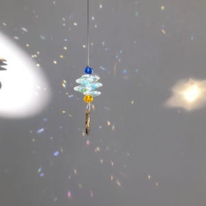 May include: A hanging crystal suncatcher with a layered design. The suncatcher features clear, iridescent crystals, blue and yellow beads, and a small gold-colored charm. The crystals are suspended from a thin wire.