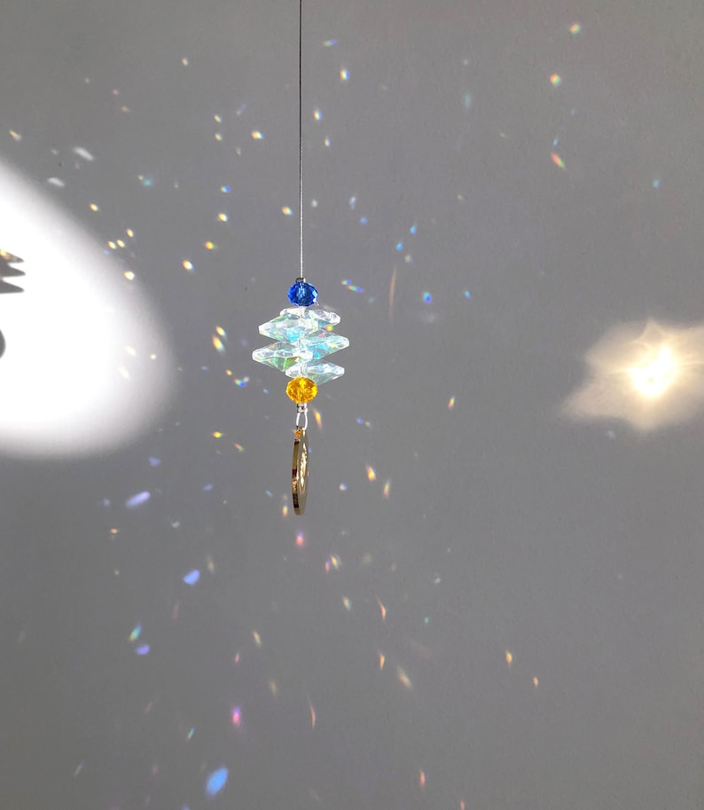 May include: A hanging crystal suncatcher with a layered design. The suncatcher features clear, iridescent crystals, blue and yellow beads, and a small gold-colored charm. The crystals are suspended from a thin wire.