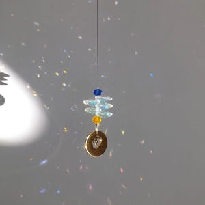 May include: A hanging suncatcher with a gold-colored charm featuring baby footprints. The suncatcher has blue, yellow, and clear crystal beads. The beads are strung on a thin wire, creating a decorative and reflective piece.