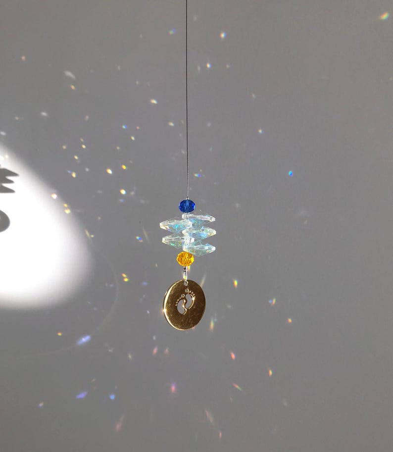 May include: A hanging suncatcher with a gold-colored charm featuring baby footprints. The suncatcher has blue, yellow, and clear crystal beads. The beads are strung on a thin wire, creating a decorative and reflective piece.