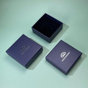 May include: Three navy blue square boxes, two closed and one open, arranged on a teal surface. The closed boxes have a silver logo and the text "NEWMERRY". The open box reveals a black interior, suggesting a jewelry or gift box.