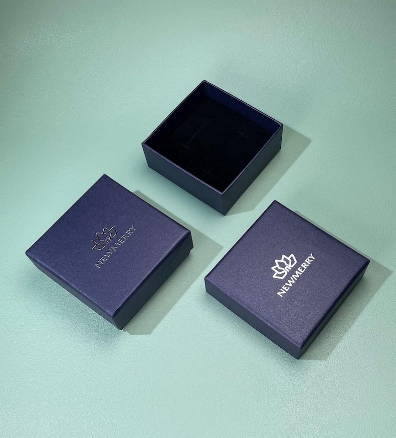 May include: Three navy blue square boxes, two closed and one open, arranged on a teal surface. The closed boxes have a silver logo and the text "NEWMERRY". The open box reveals a black interior, suggesting a jewelry or gift box.