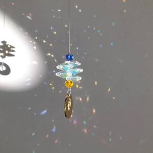 May include: A hanging crystal suncatcher with a blue and yellow bead design. The suncatcher has a gold-colored charm at the bottom and is suspended by a thin wire. The light reflects off the crystals, creating a rainbow effect.