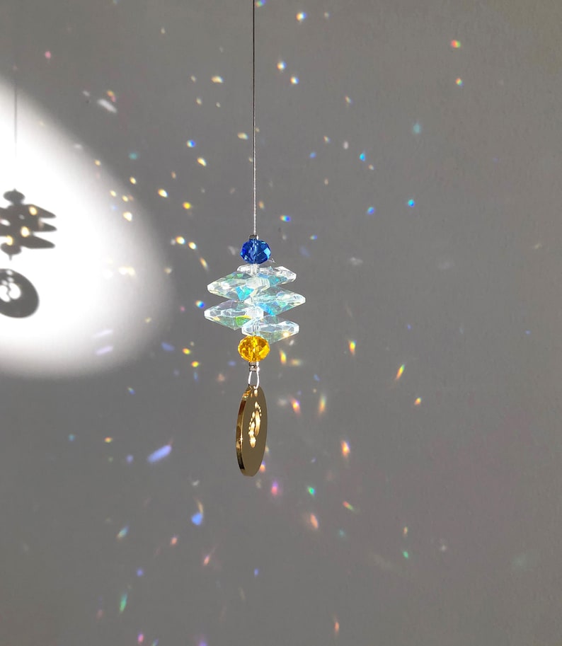 May include: A hanging crystal suncatcher with a blue and yellow bead design. The suncatcher has a gold-colored charm at the bottom and is suspended by a thin wire. The light reflects off the crystals, creating a rainbow effect.