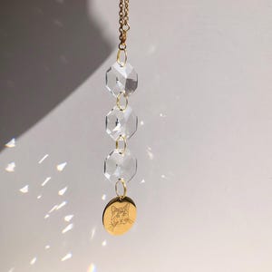 May include: A clear crystal suncatcher with three faceted crystal beads and a gold-colored chain. A small, round wooden charm with a cat face is at the bottom. The suncatcher hangs against a white background.
