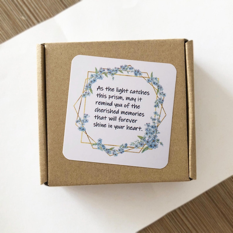 May include: A small, square, brown cardboard box with a white sticker on top. The sticker features a gold geometric design with blue floral accents and text that reads, "As the light catches this prism, may it remind you of the cherished memories that will forever shine in your heart."
