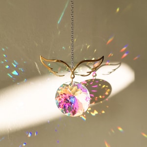 Peut inclure: Un attrape-soleil en forme d'aile d'ange en métal doré avec une boule de cristal transparente suspendue. La boule de cristal est irisée et projette des arcs-en-ciel sur le mur.