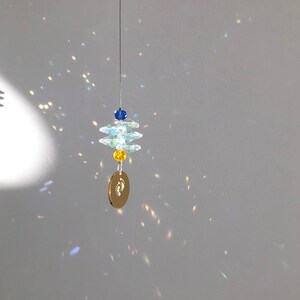 May include: A hanging suncatcher with a gold-colored oval pendant featuring a baby footprint design. The suncatcher has clear, faceted crystal elements, blue and yellow beads, and a thin silver wire for hanging. The light refracts through the crystals.