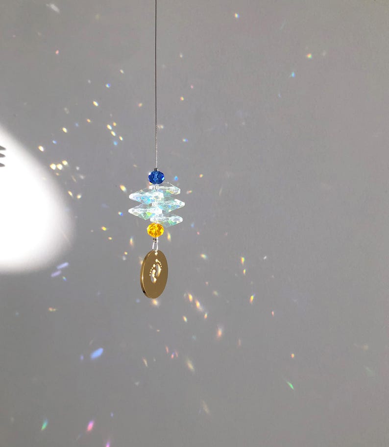 May include: A hanging suncatcher with a gold-colored oval pendant featuring a baby footprint design. The suncatcher has clear, faceted crystal elements, blue and yellow beads, and a thin silver wire for hanging. The light refracts through the crystals.