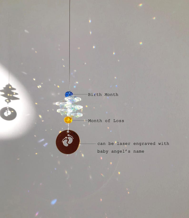 May include: A hanging memorial ornament with a clear crystal design, blue and yellow beads, and a circular copper charm with baby footprints. Text on the charm reads, "can be laser engraved with baby angel's name."