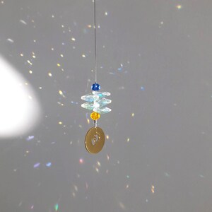 May include: A hanging suncatcher with a gold-colored disc featuring baby footprints. The suncatcher has clear, faceted crystals and blue and yellow beads. The crystals and beads are strung on a thin wire.