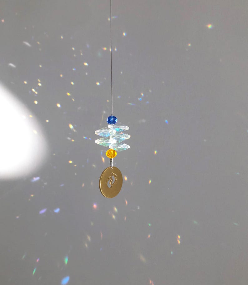 May include: A hanging suncatcher with a gold-colored disc featuring baby footprints. The suncatcher has clear, faceted crystals and blue and yellow beads. The crystals and beads are strung on a thin wire.