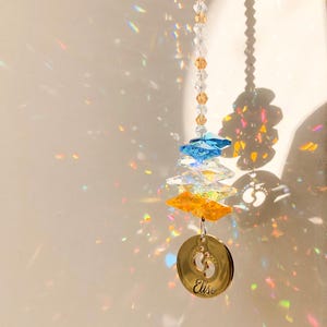 May include: A crystal suncatcher with a gold-colored charm featuring a footprint design and the name "Elise" engraved on it. The suncatcher has blue, clear, and yellow crystals.