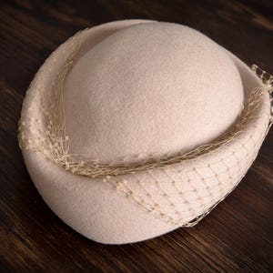 以下が含まれることがあります： 繊細なネットトリムが付いた薄いベージュのフェルトハット。帽子は丸いクラウンと広いブリムを持っています。