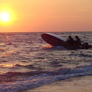 May include: A dark-colored rigid inflatable boat with an outboard motor navigating choppy ocean waves during a golden sunset. The vessel is angled upward as it cuts through the water, with the bright sun low on the horizon casting light.