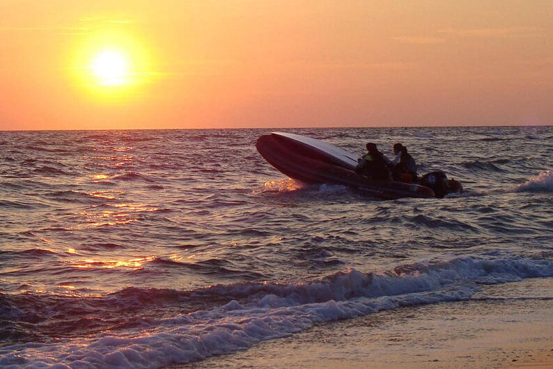 May include: A dark-colored rigid inflatable boat with an outboard motor navigating choppy ocean waves during a golden sunset. The vessel is angled upward as it cuts through the water, with the bright sun low on the horizon casting light.
