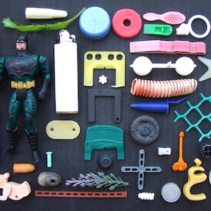 Puede incluir: Una colección de pequeños objetos de plástico variados que incluye una figura de acción de color verde oscuro y negro, un encendedor Bic azul, un encendedor blanco, una moneda dorada, una planta de plástico verde, una pieza en forma de estrella, un neumático negro y varias formas.