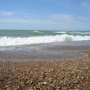 May include: A close-up of a pebbled beach with a wave crashing on the shore. The water is a light blue-green color and the sky is a clear blue.