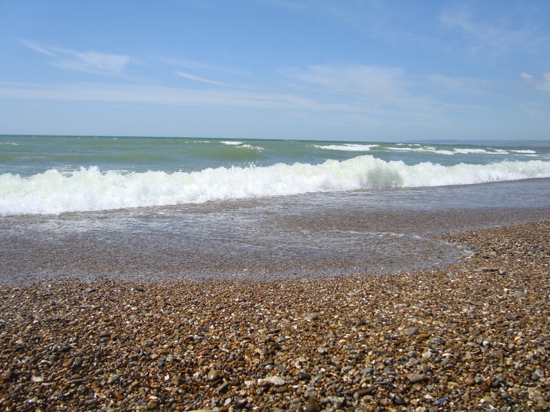 May include: A close-up of a pebbled beach with a wave crashing on the shore. The water is a light blue-green color and the sky is a clear blue.