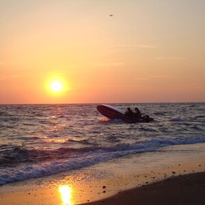 May include: A silhouette of a small boat with two people on board, speeding across the water towards the shore. The sun is setting in the distance, casting a warm orange glow over the scene.